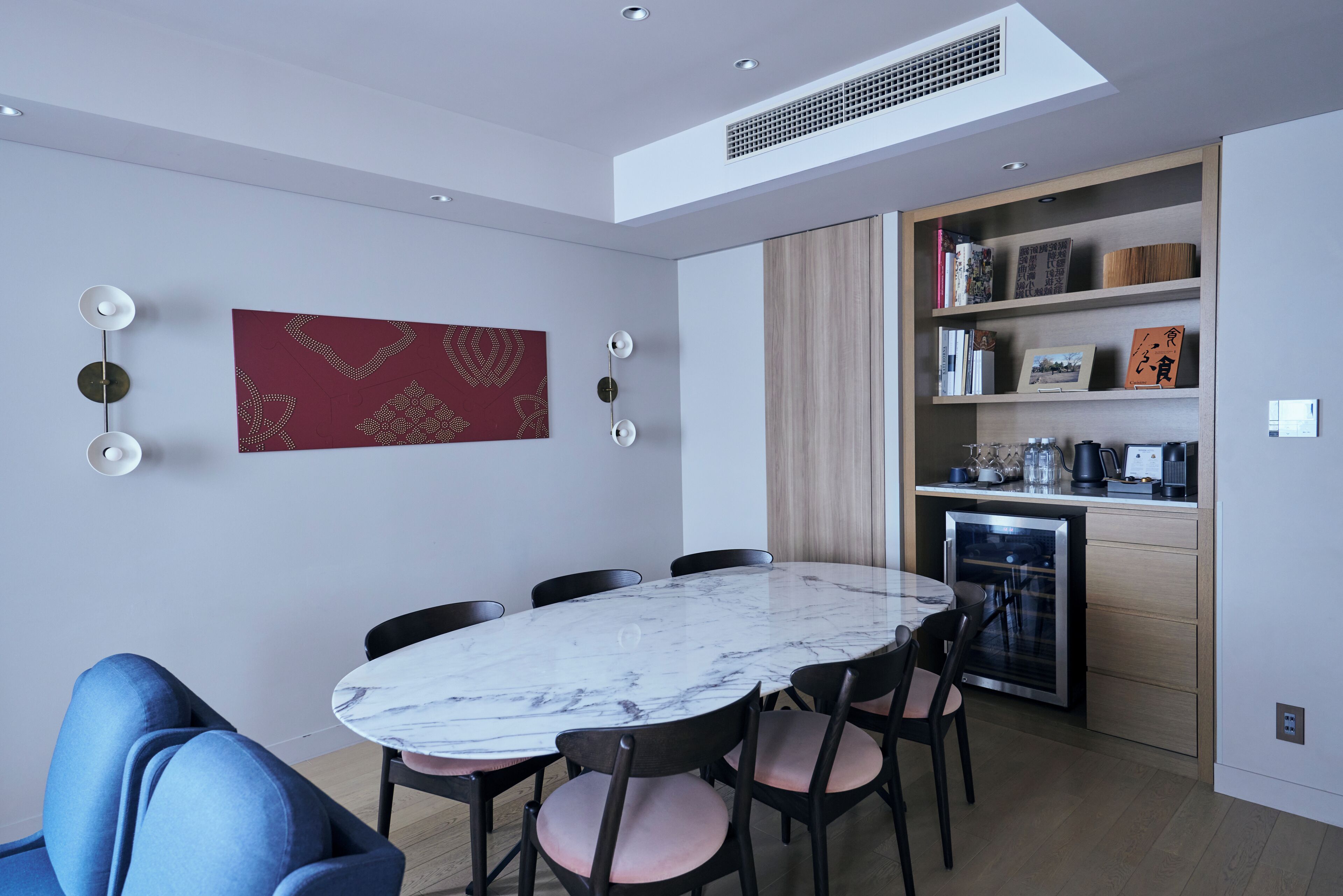 A modern dining area with a marble table and chairs, a red patterned wall hanging, and a built-in kitchen with shelves.