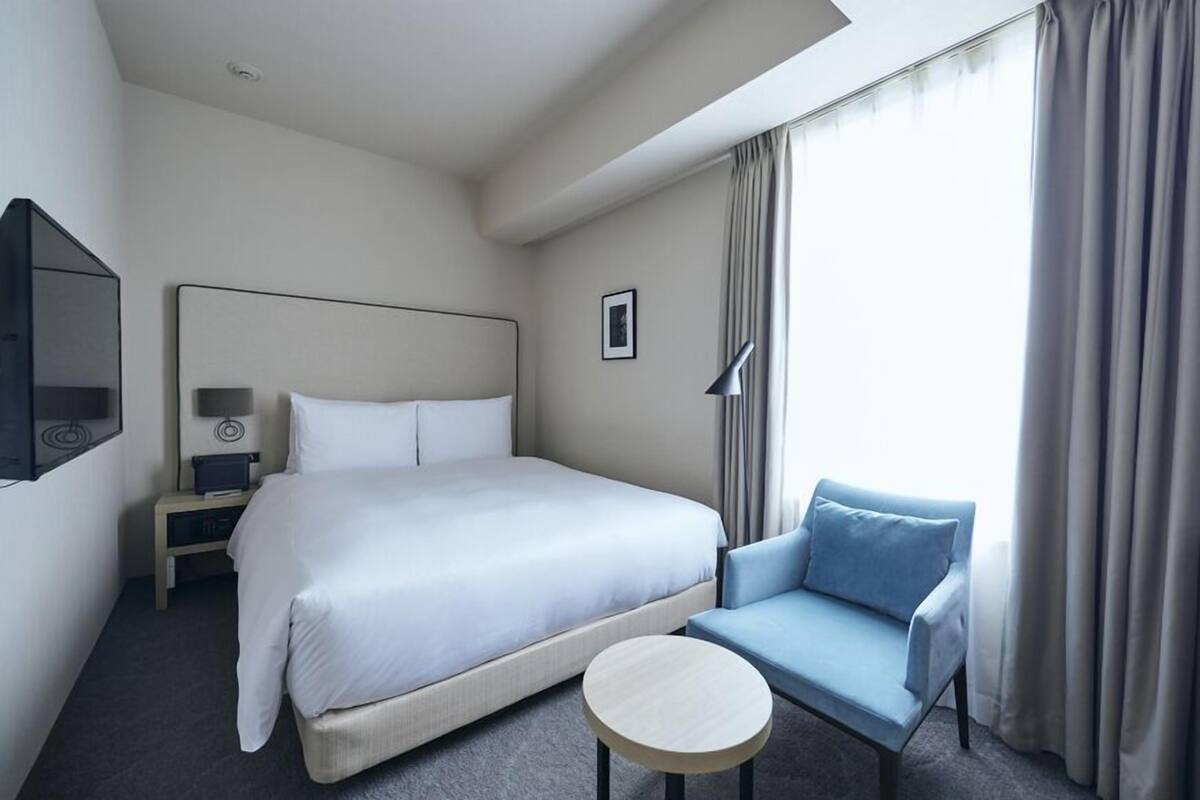 A hotel room with a bed, a chair, a small table, a nightstand, a lamp, and a picture on the wall.