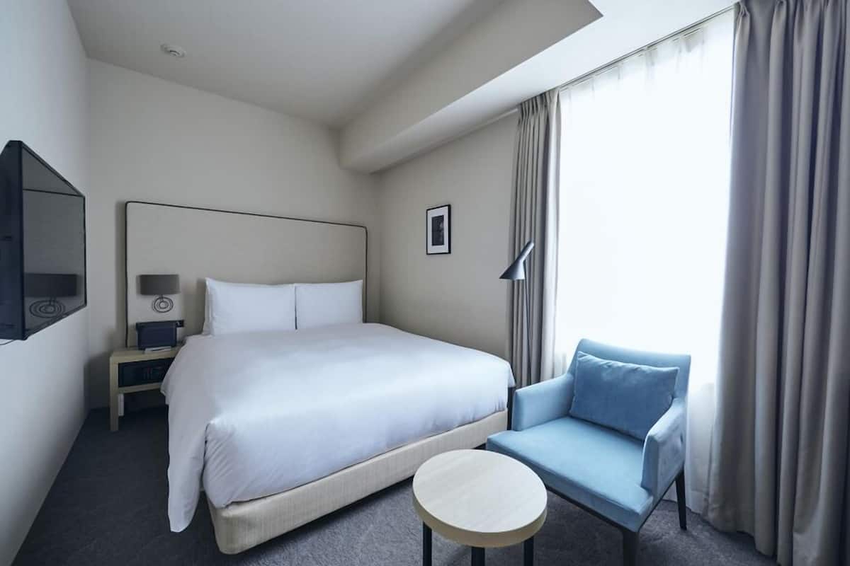 Habitación de hotel con cama, una silla, una mesita, una mesita de noche, una lámpara y un cuadro en la pared.