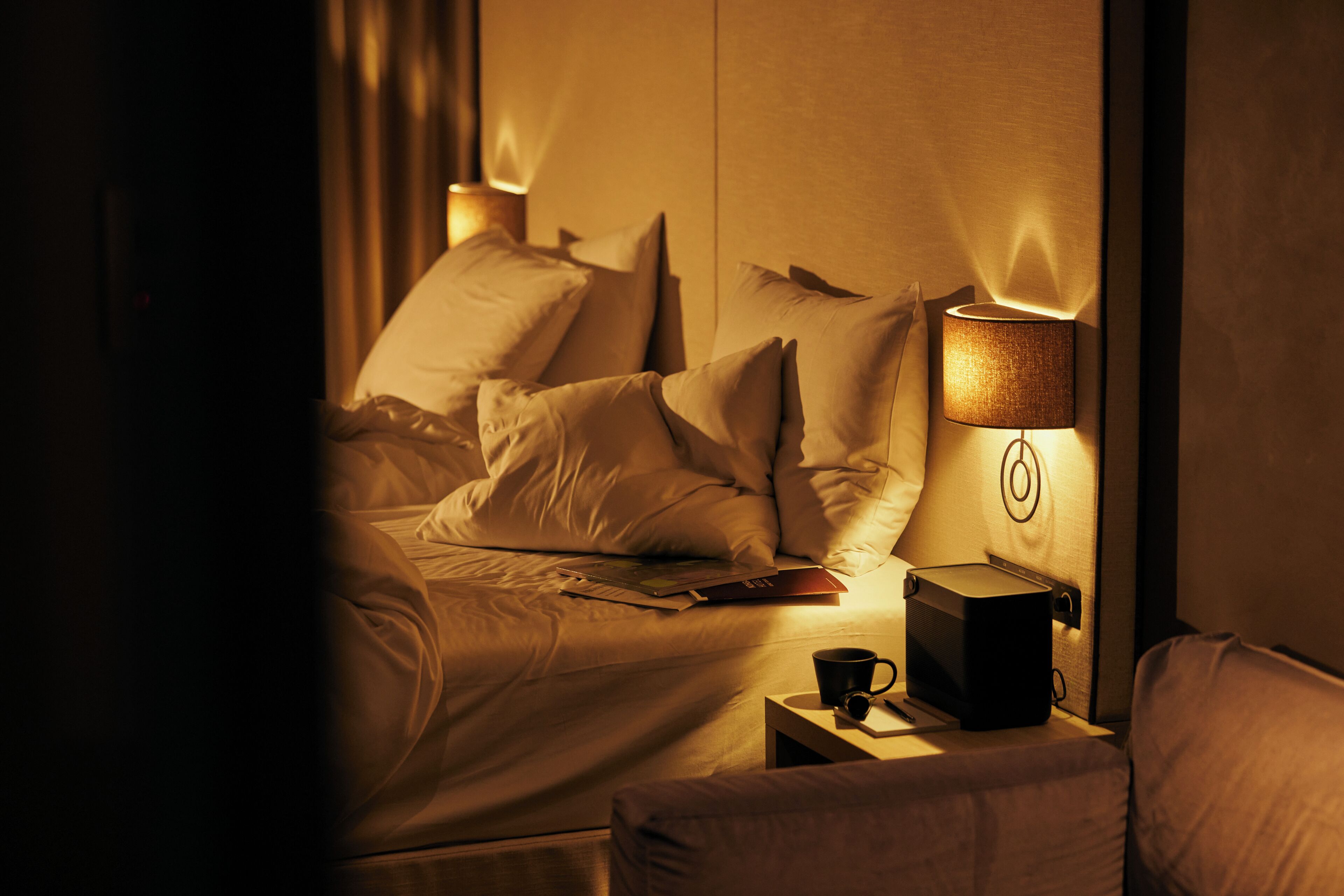 Hotellihuone, jossa on sänky, yöpöytä lampun kanssa, kuppi ja kirja.