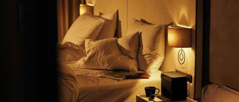 A hotel room with a bed, a bedside table with a lamp, a cup, and a book.