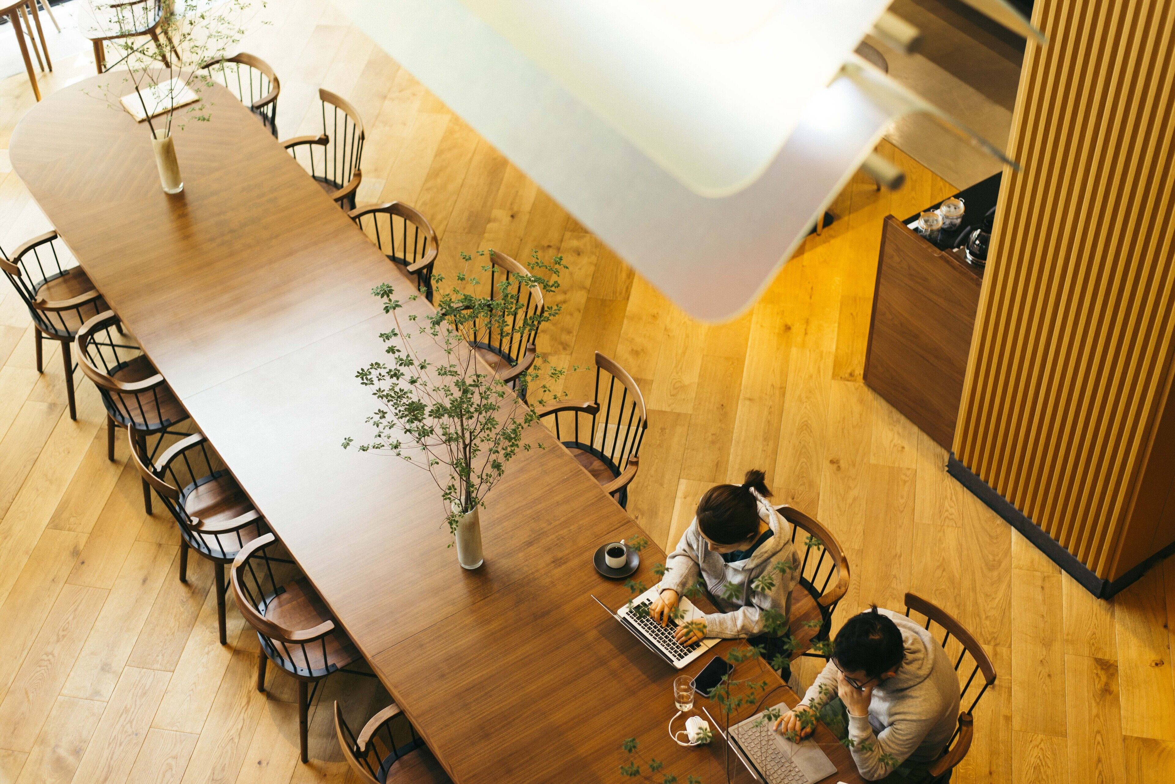 A long wooden dining table with chairs, a vase with greenery, and two people working on laptops.