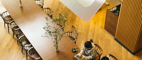 A long wooden dining table with chairs, a vase with greenery, and two people working on laptops.