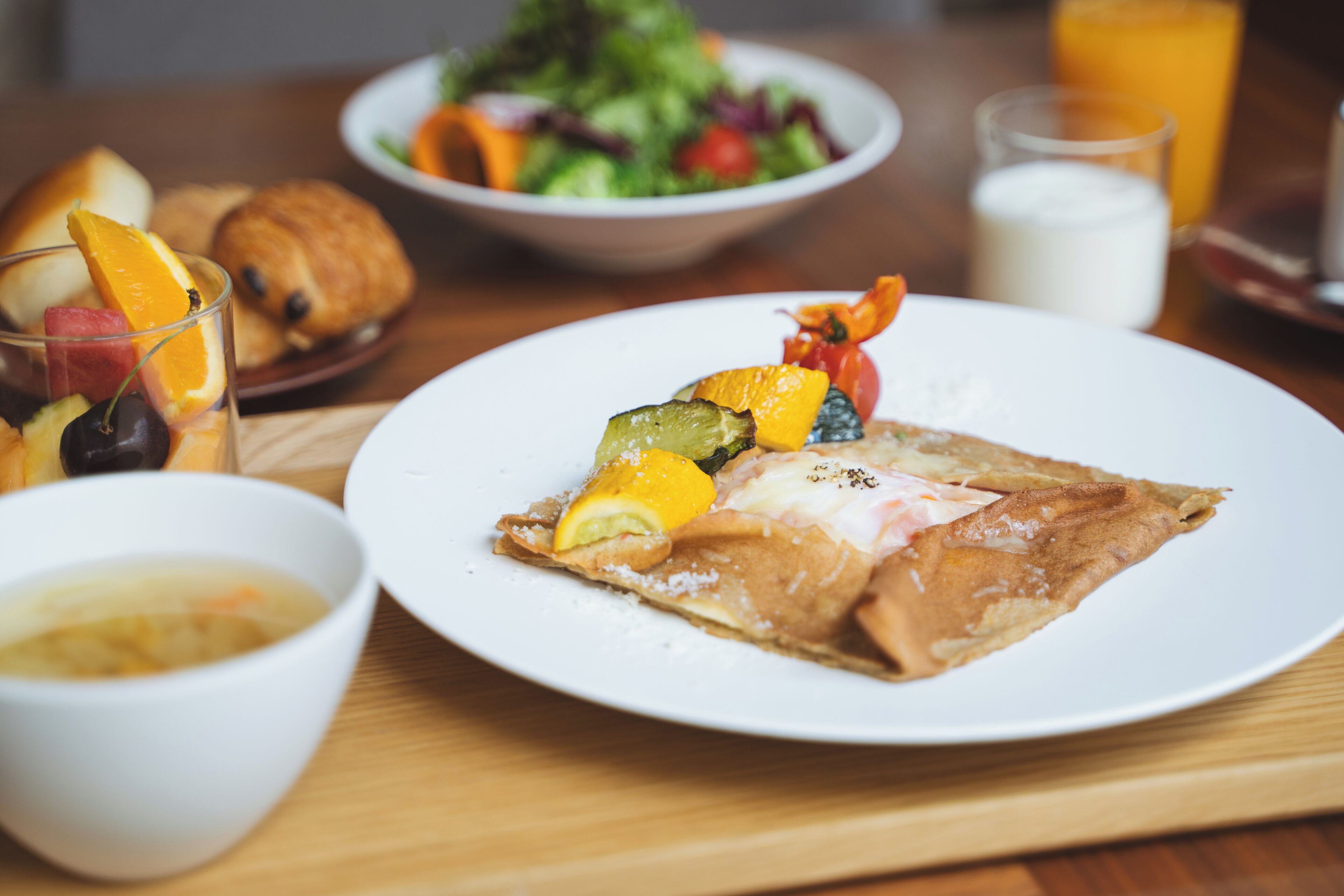 A breakfast setting with a plate of food, a bowl of soup, a croissant, a glass of orange juice, and a glass of milk.