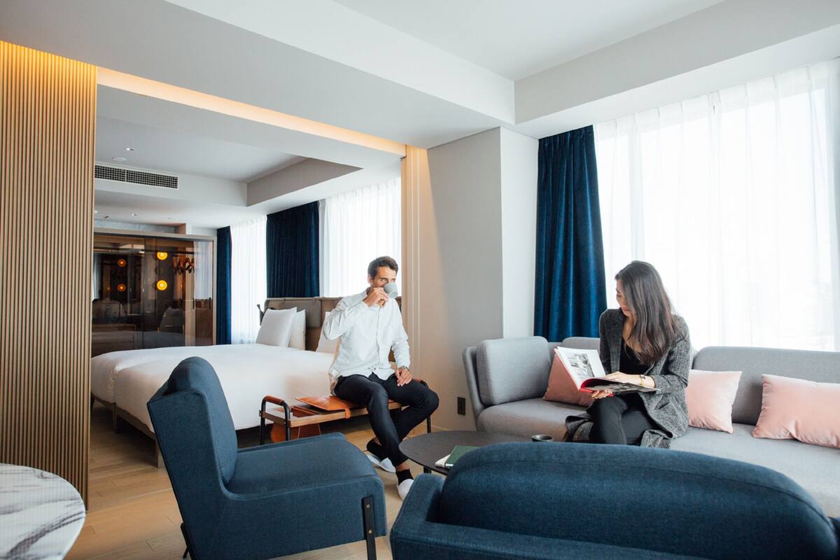 A modern hotel room with a large bed, two seating areas, and a view of the city through large windows.