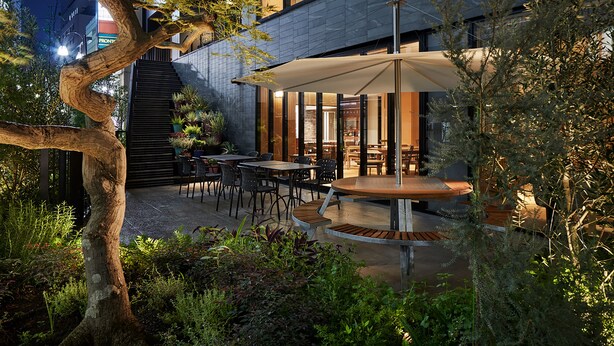 Un patio al aire libre con mesas y sillas, rodeado de vegetación y una escalera que lleva a un nivel superior.
