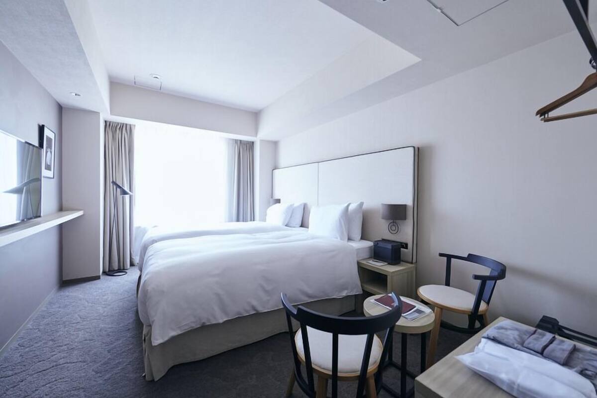 A hotel room with a large bed, a desk with a chair, a bedside table, and a window with curtains.