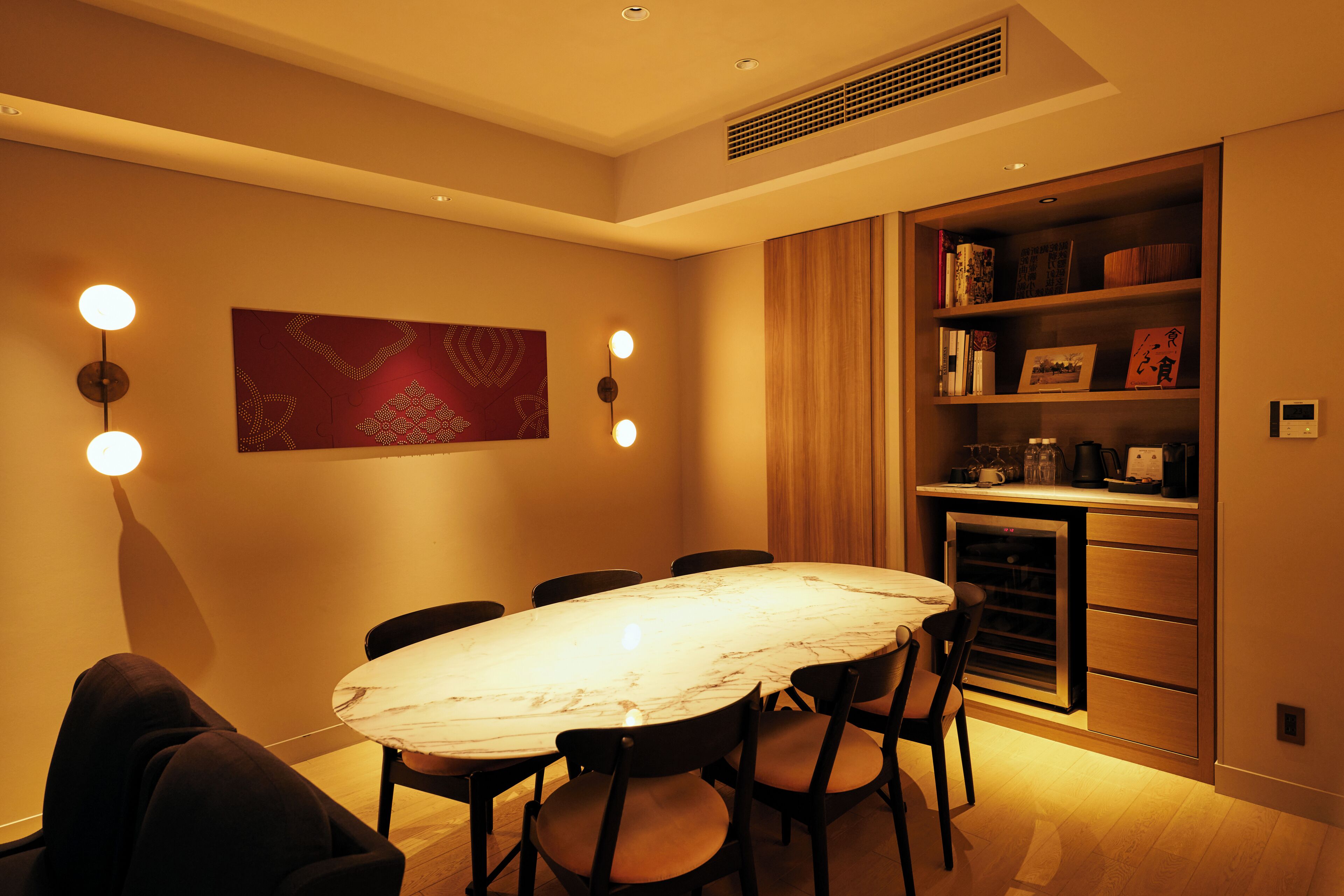 A dining area with a marble table, chairs, wall-mounted lights, and a bookshelf with books and decorative items.