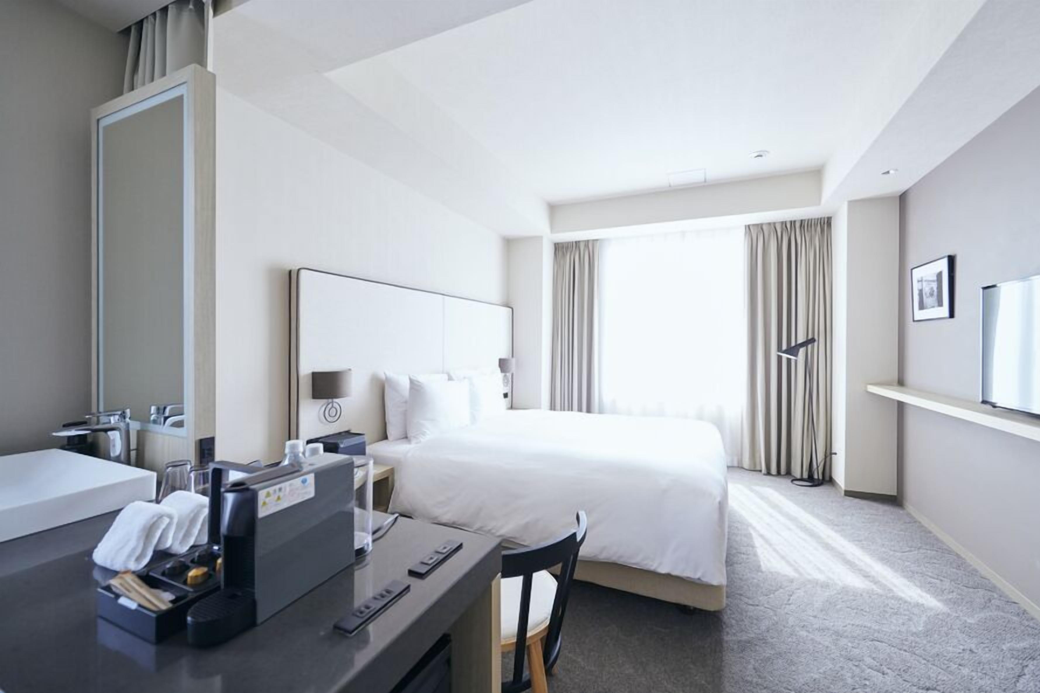 A hotel room with a bed, a desk with a computer, a mirror, and a window with curtains.