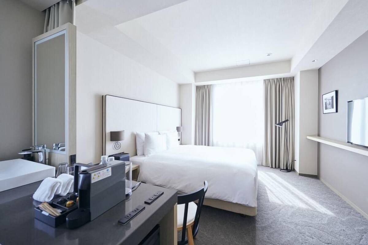A hotel room with a bed, a desk with a computer, a mirror, and a window with curtains.