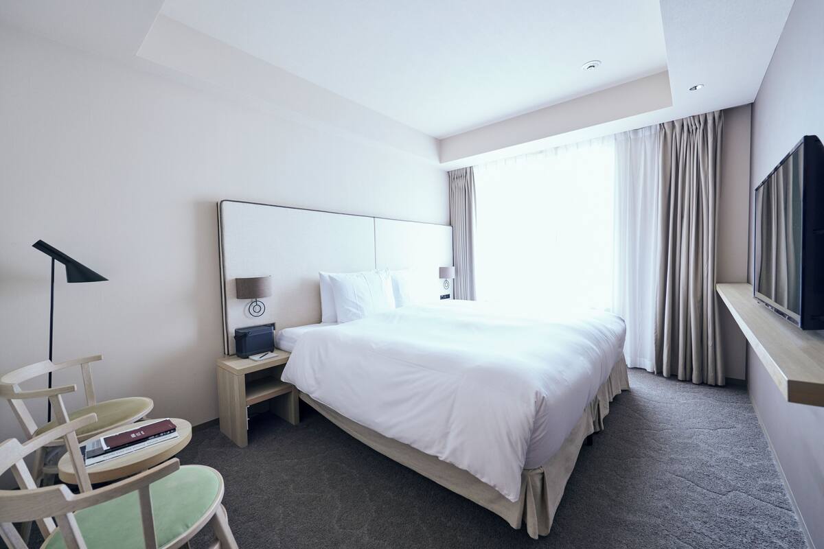 A hotel room with a large bed, a desk with a lamp, a chair, and a television on a stand.