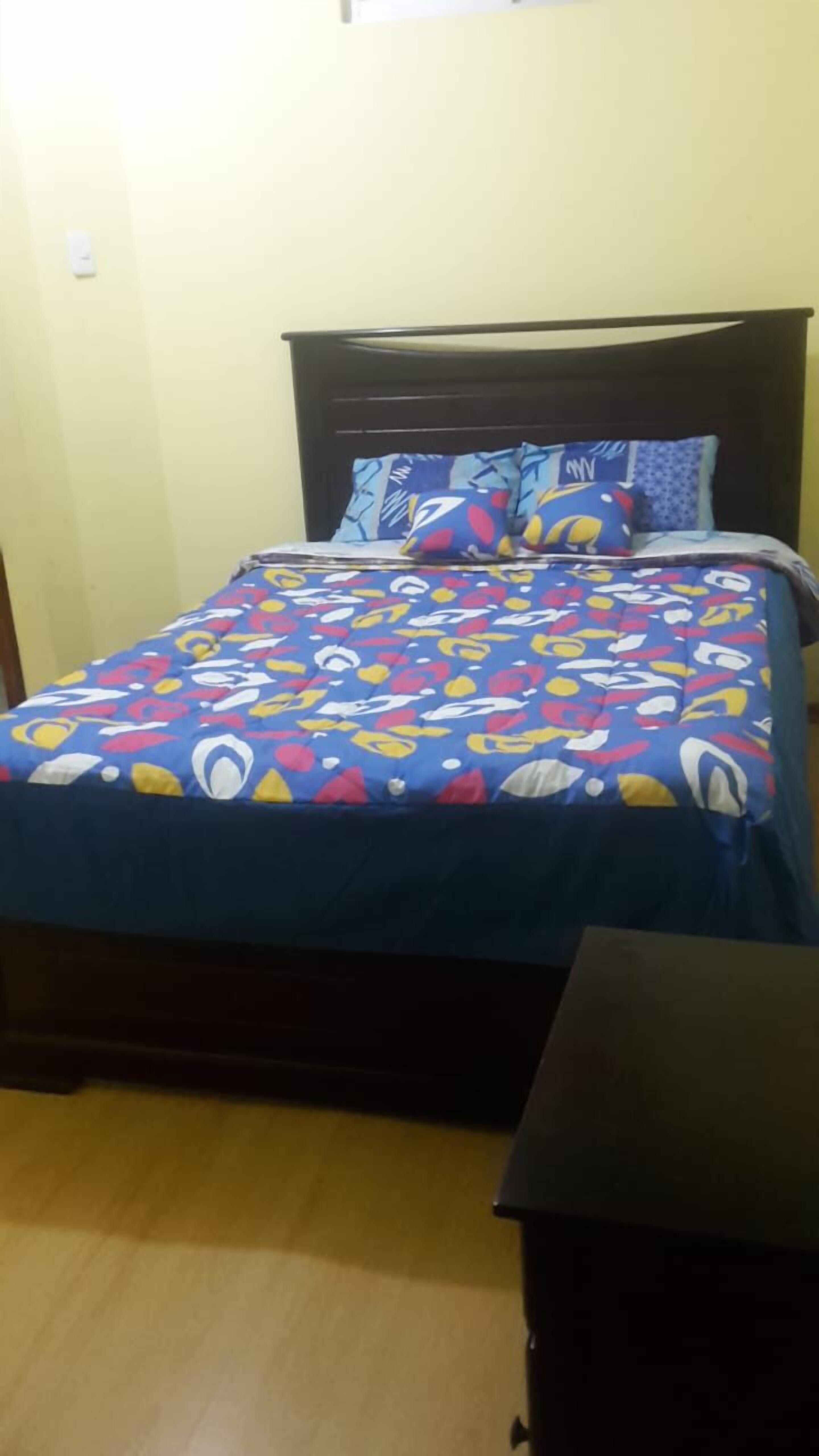 A bed with a colorful patterned bedspread and matching pillows, a dark wooden headboard, and a small bedside table.