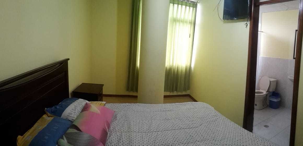A bedroom with a bed, a nightstand, a window with curtains, a television on the wall, and a bathroom visible through an open door.