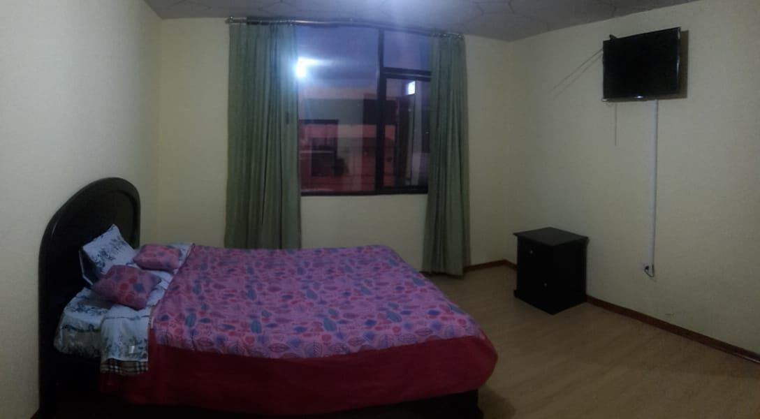 Un dormitorio con una cama, un televisor montado en la pared y una ventana con cortinas.