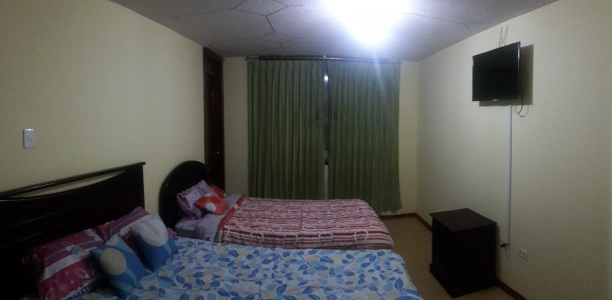 Un dormitorio con cama, televisor, cortina y una pequeña mesa.