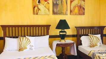 A hotel room with two beds, each with a wooden headboard and white bedding. There is a small table with a green lamp between the beds. The walls are yellow, and there are three paintings above the beds.