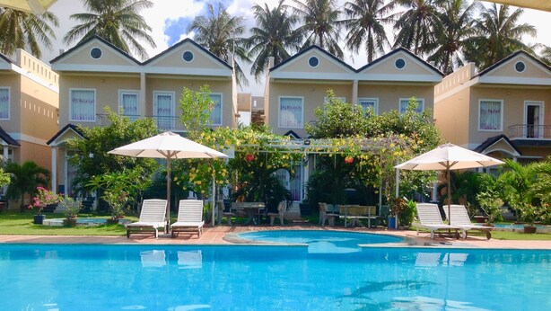 A swimming pool with lounge chairs and umbrellas, surrounded by palm trees and a building with multiple windows. - Areca Resort Cay Cau (Mui Ne)