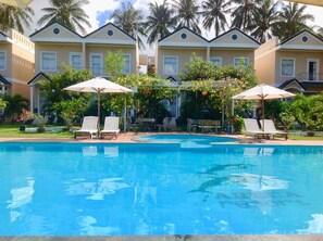 A swimming pool with lounge chairs and umbrellas, surrounded by palm trees and a building with multiple windows.