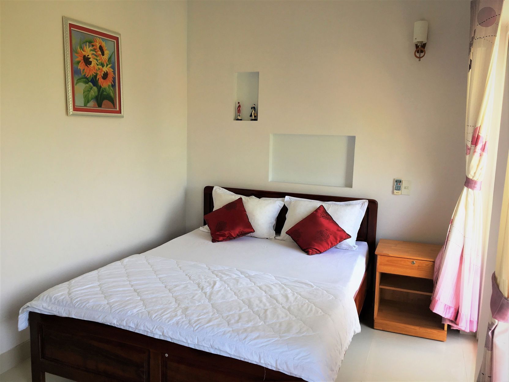 A bed with white bedding and red pillows, a nightstand with a phone, a painting of sunflowers, and a curtain.