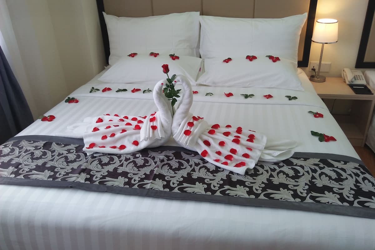 A neatly made bed with white linens, red rose petals, and a swan-shaped towel arrangement.