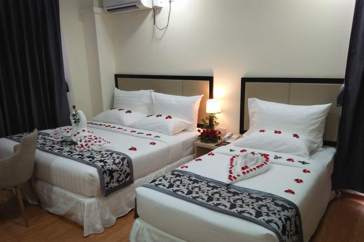 A hotel room with two beds, each with white linens and red rose petals arranged on the beds. There is a bedside table with a lamp and a phone. The room has a wooden floor and a window with curtains.