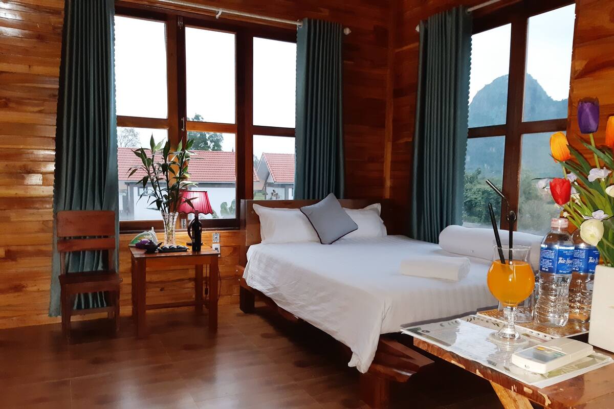 A wooden room with a bed, a desk, and a chair. There are curtains, a lamp, and a vase with flowers.