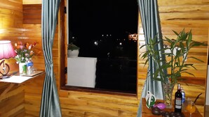 A wooden cabin room with a window, curtains, a table with a lamp, a vase with flowers, a potted plant, a bottle of wine, and a glass of orange juice. - Diep Le Homestay (Phong Nha)