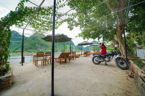 A covered outdoor seating area with wooden chairs and tables, a motorcycle, and a view of a river and hills in the background. - Diep Le Homestay (Phong Nha)