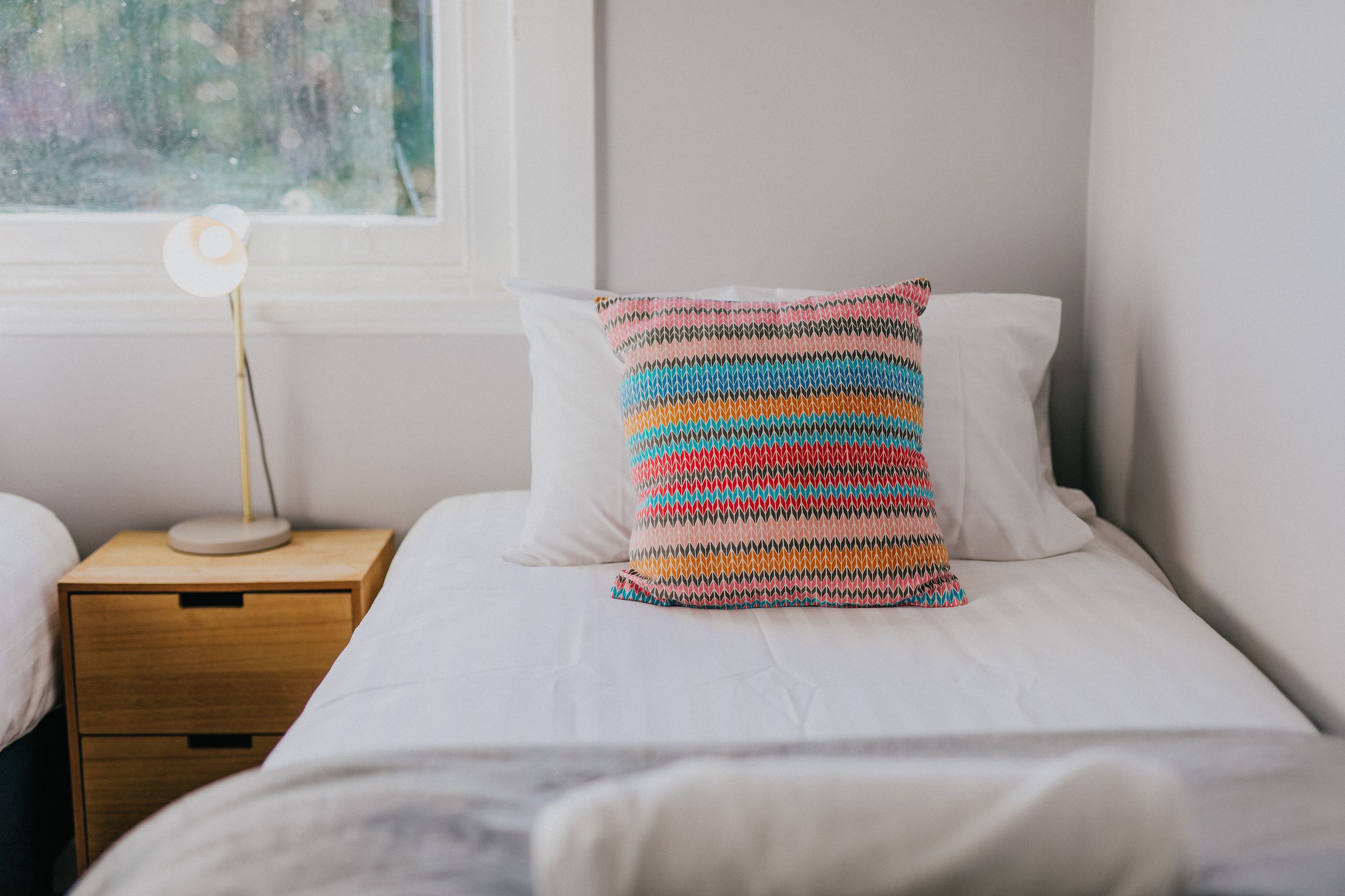 Een opgemaakt bed met een kleurrijk gestreept kussen en een nachtkastje met een lamp.