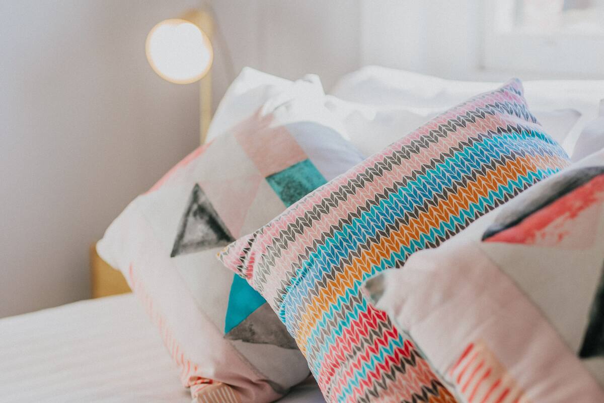 A bed with multiple pillows, a bedside lamp, and a window in the background.