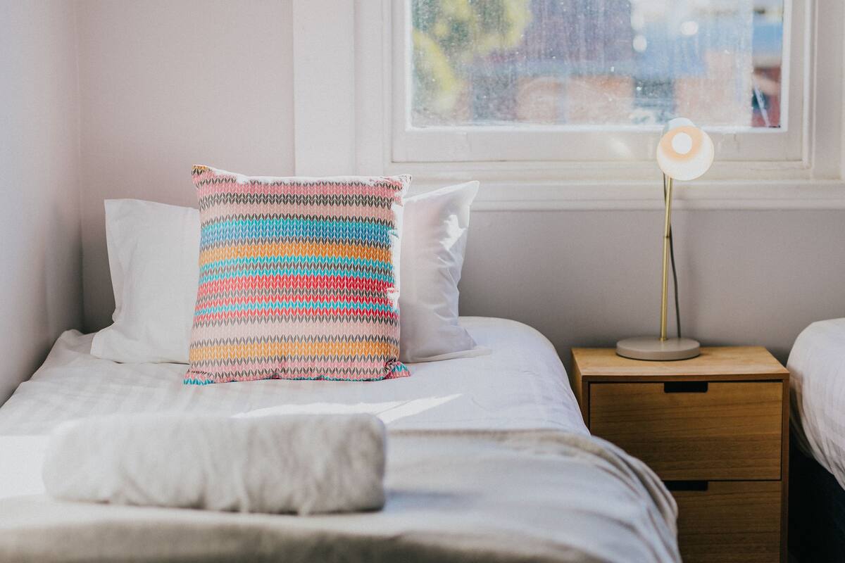 A neatly made bed with a colorful striped pillow and a white pillow, a bedside lamp, and a window with a view of buildings.