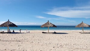 A beach with three thatched umbrellas, a sandy shore, and the ocean in the background. - Mario Hotel and Cafe (Tambolaka)