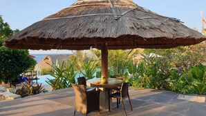 A thatched umbrella structure with a table and chairs underneath, surrounded by lush greenery and a pool in the background. - Mario Hotel and Cafe (Tambolaka)