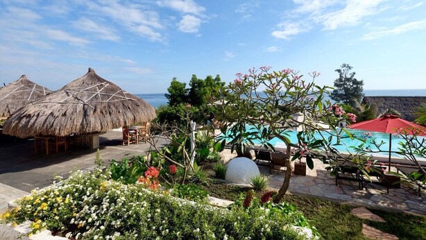 A poolside area with thatched umbrellas, lounge chairs, and a view of the sea. - Mario Hotel and Cafe (Tambolaka)
