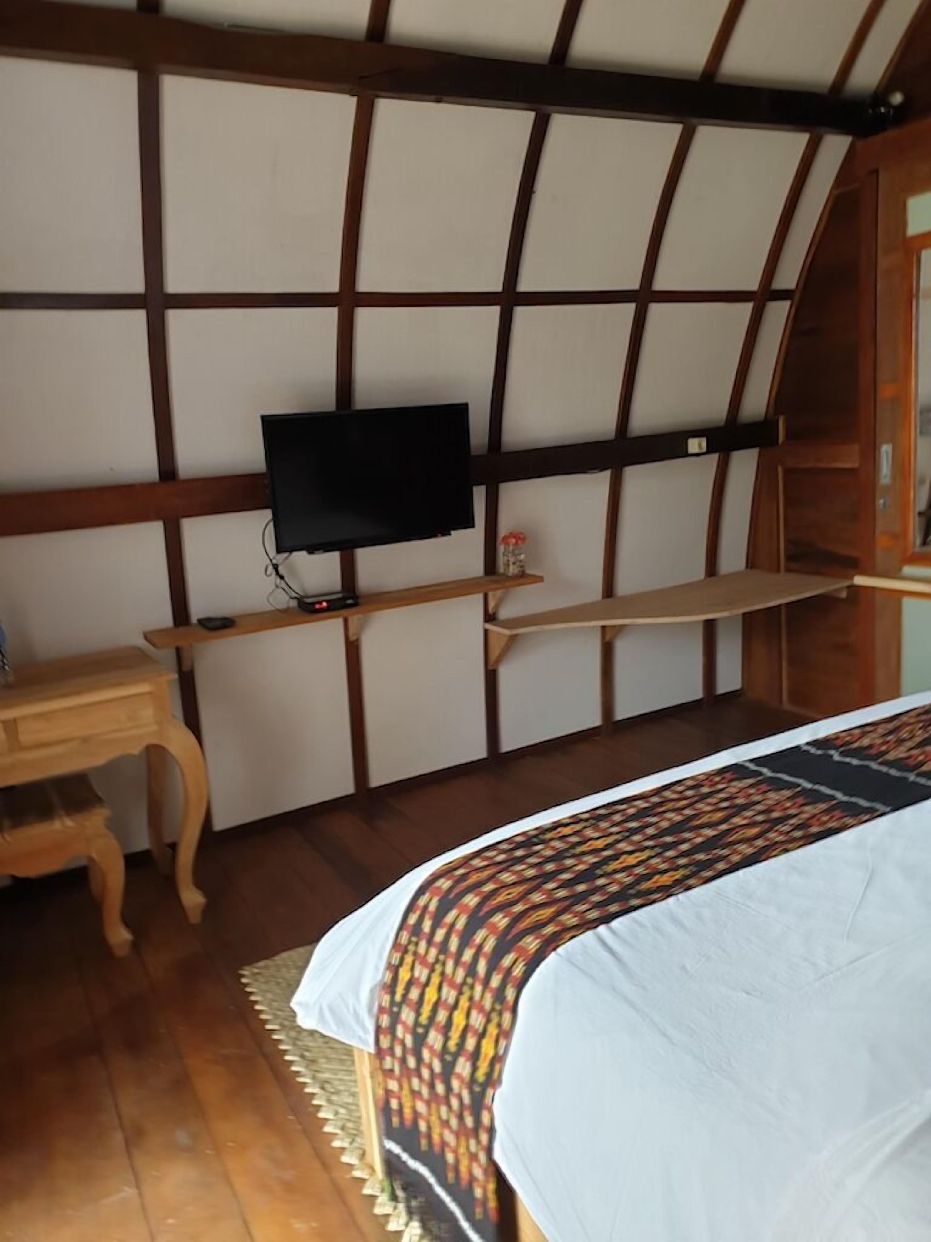 A bedroom with a bed, a TV mounted on a wooden wall, a small wooden table, and a shelf.