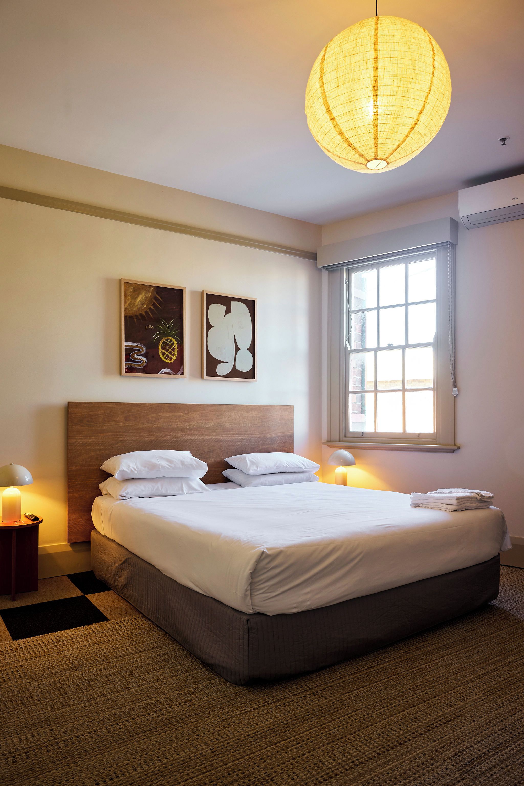 A bedroom with a large bed, two bedside lamps, a window with blinds, and two wall art pieces.