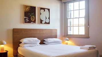 A bedroom with a large bed, two bedside lamps, a window with blinds, and two wall art pieces.