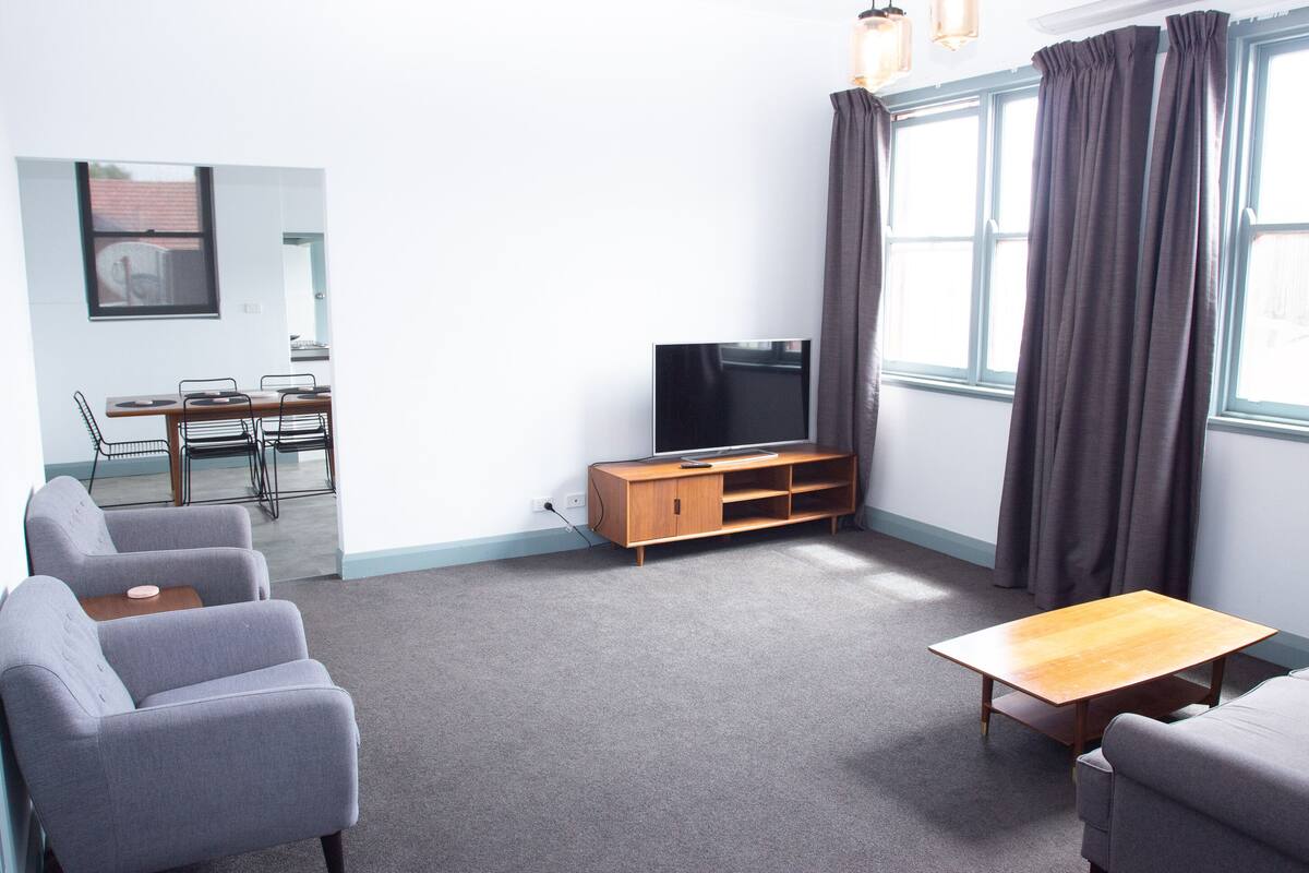A modern living room with grey sofas, a wooden coffee table, a TV on a wooden stand, and large windows with curtains.