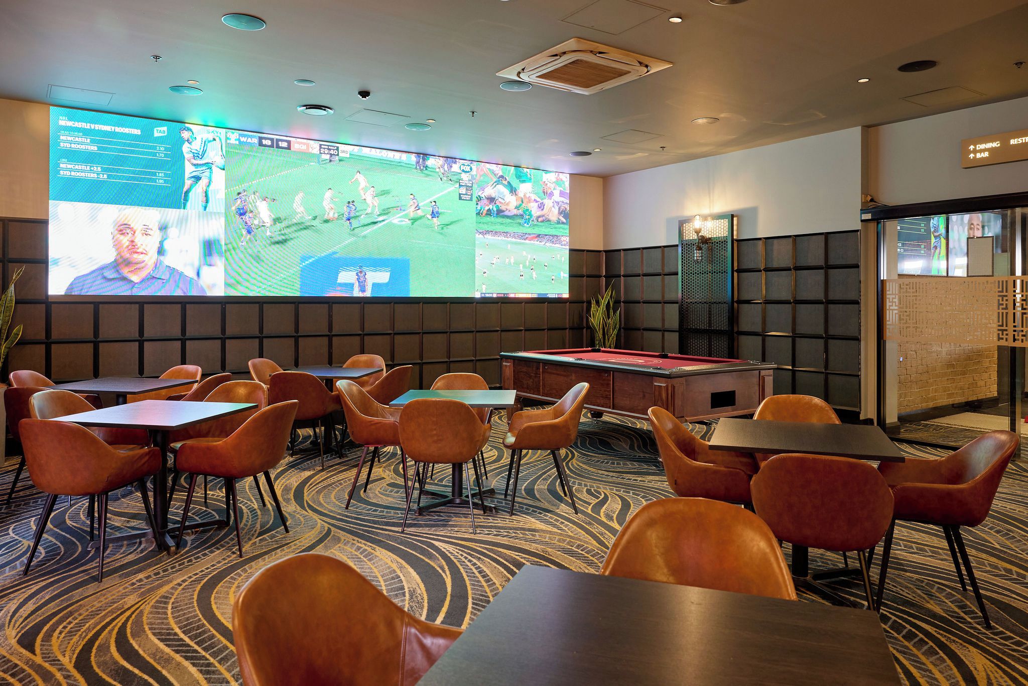 Una habitación con mesa de billar, varios conjuntos de asientos y una pantalla grande mostrando un evento deportivo.