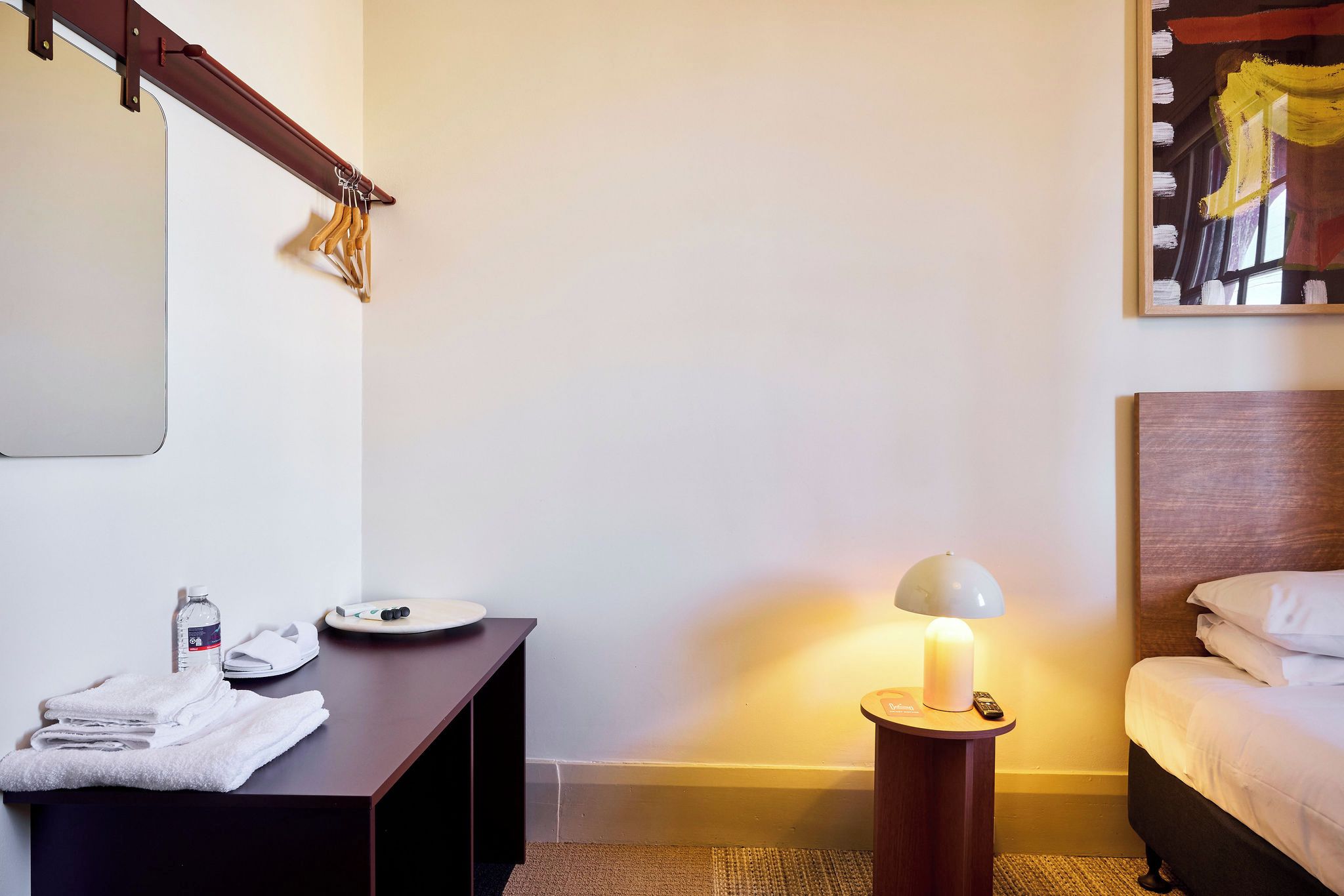 A hotel room with a bed, a bedside table with a lamp, a towel stack, and a wall-mounted towel rack.