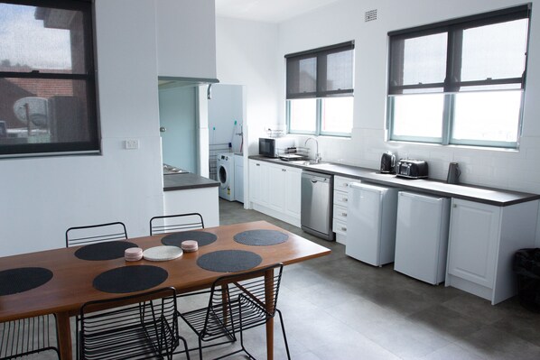 A modern kitchen with white cabinetry, a dining table with black placemats, and a window with blinds. - Oasis on Beamish Hotel (Campsie)