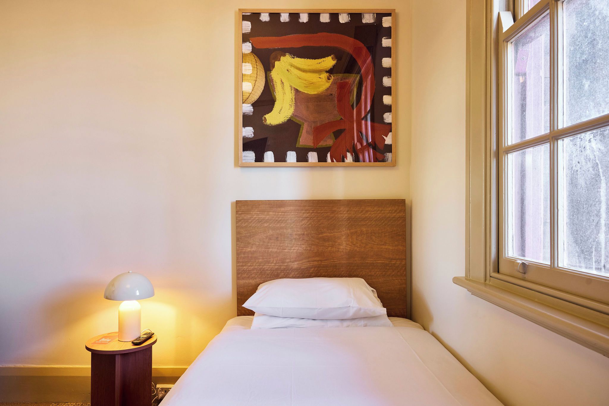 A hotel room with a bed, a nightstand with a lamp, a painting on the wall, and a window.