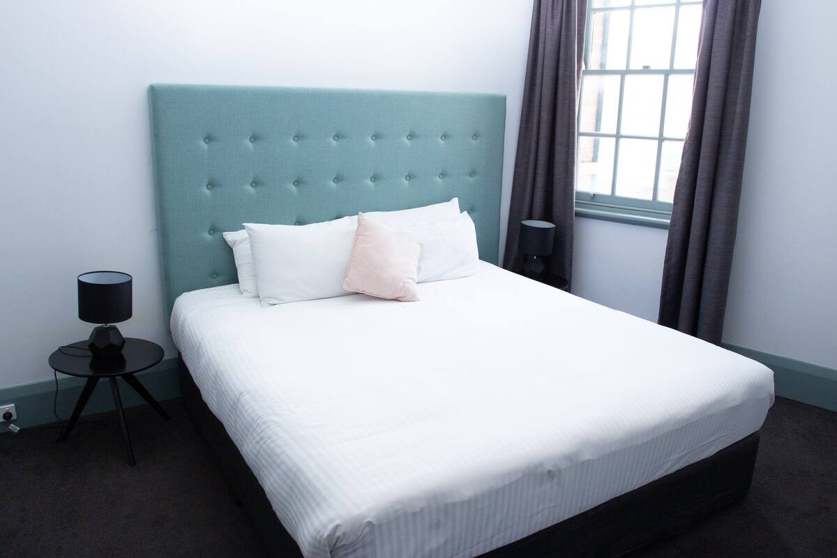 A neatly made bed with a tufted headboard, a bedside lamp, and a window with curtains.