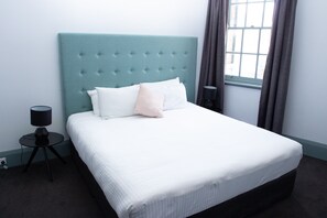 A neatly made bed with a tufted headboard, a bedside lamp, and a window with curtains. - Oasis on Beamish Hotel (Campsie)