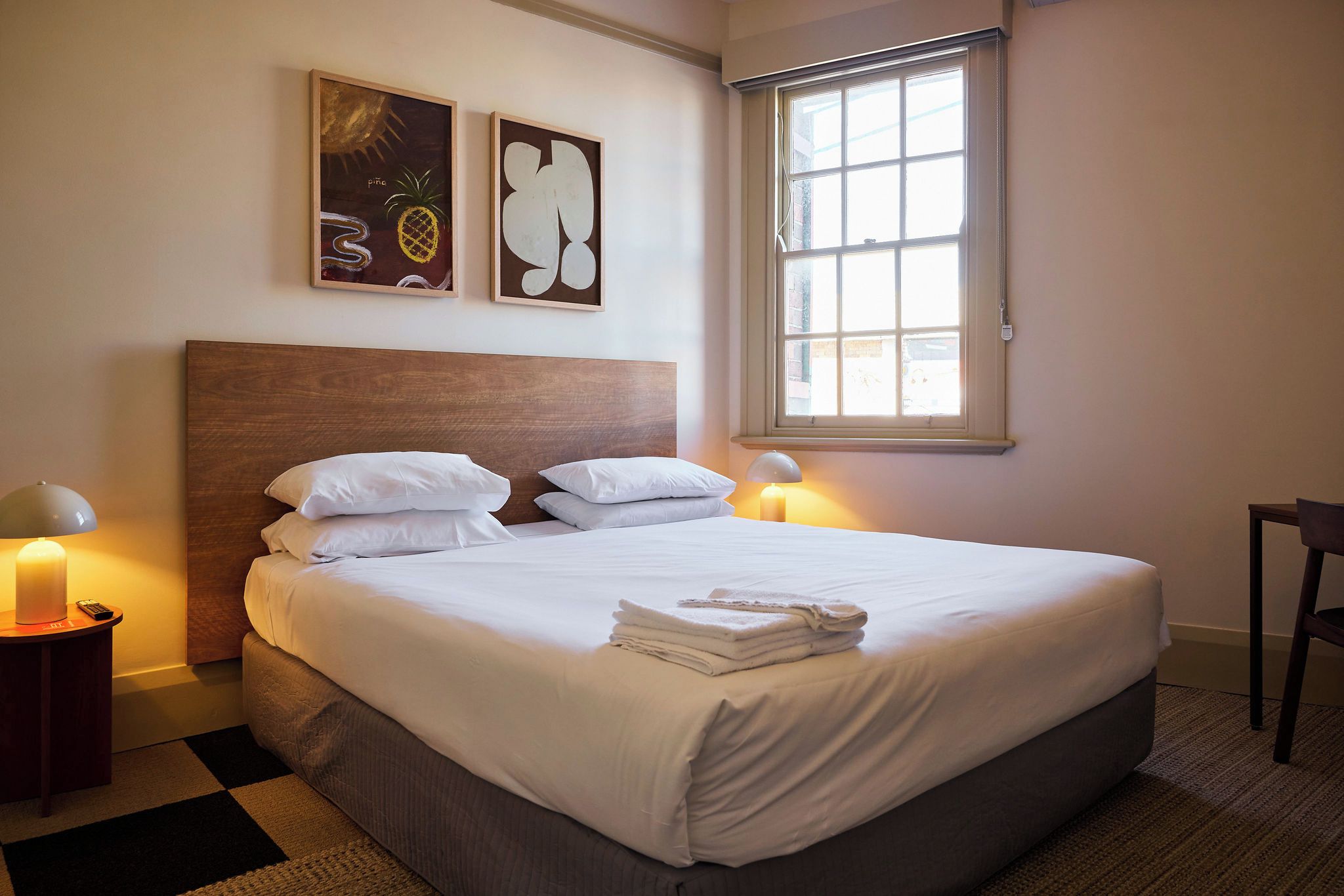 A neatly made bed with white linens and a folded towel, a wooden headboard, a window with daylight, and two bedside lamps.