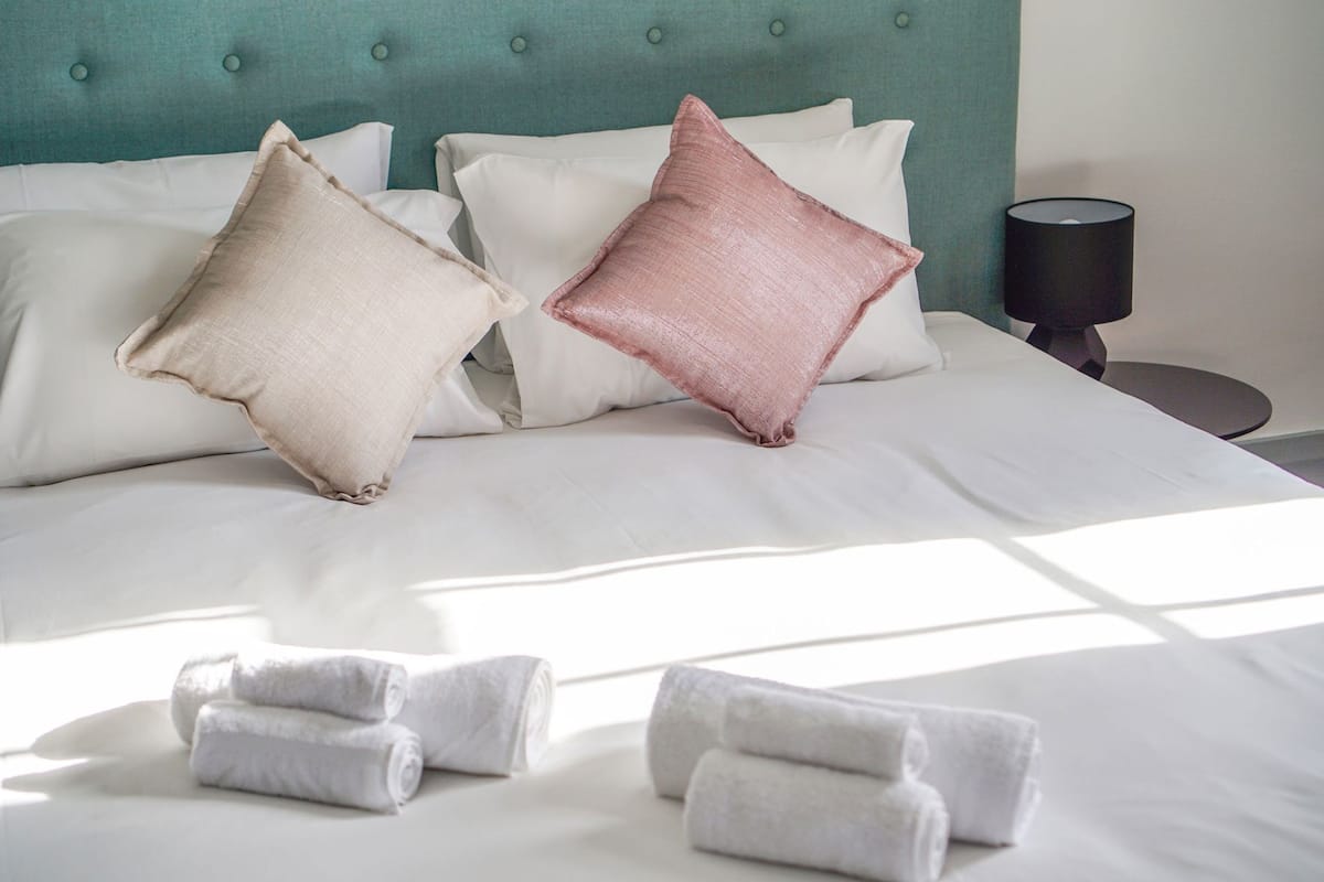 A neatly made bed with white and pink pillows, a teal headboard, and rolled towels on the bed.