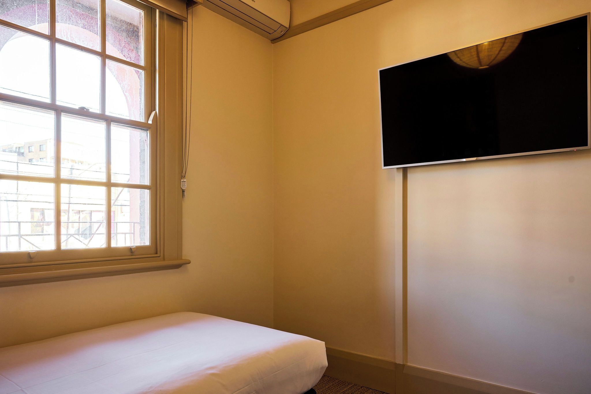 Ein kleines Zimmer mit einem Einzelbett, Fenster, einem an der Wand befestigten Fernseher und einer Steckdose.