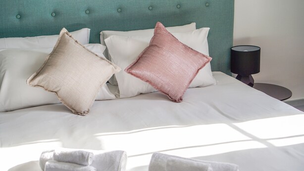 Una cama bien hecha con sábanas blancas y dos almohadas, un cabecero color turquesa y una lámpara de noche.