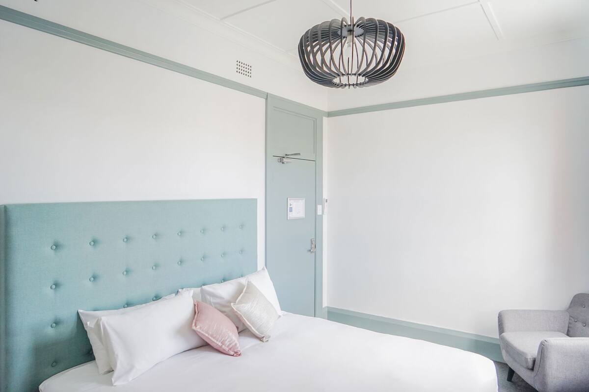 A bedroom with a bed, a chair, a light blue wall, a white ceiling, and a unique chandelier.