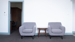 Una habitación con dos sillones grises y una pequeña mesa de madera, una puerta que da a otra estancia, y suelo cubierto de alfombra.